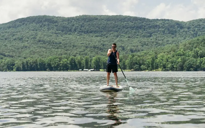 Man staat op een SUP board op een kalm meer met beboste heuvels op de achtergrond